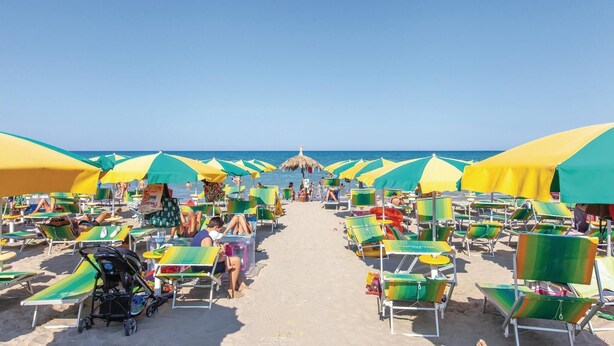Ein Strand mit Reihen von Liegestühlen und Sonnenschirmen, einem klaren Himmel und dem Meer im Hintergrund.