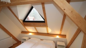 Ein Schlafzimmer mit schrägem Dach, einem Bett, Nachttischen und einem Fenster mit Blick auf ein Gebäude.