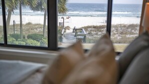 Blick auf einen Strand durch ein Fenster, mit einer Holzterrasse und sichtbaren Palmen.