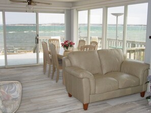 A room with a beige leather sofa, wooden chairs, a dining table with a vase of flowers, and large windows overlooking the sea. - SUMMER AUGUST & SEPTEMBER PRIVATE BEACH ON NOYAC BAY SAG HARBOR SPECTACULAR! (Sag Harbor)