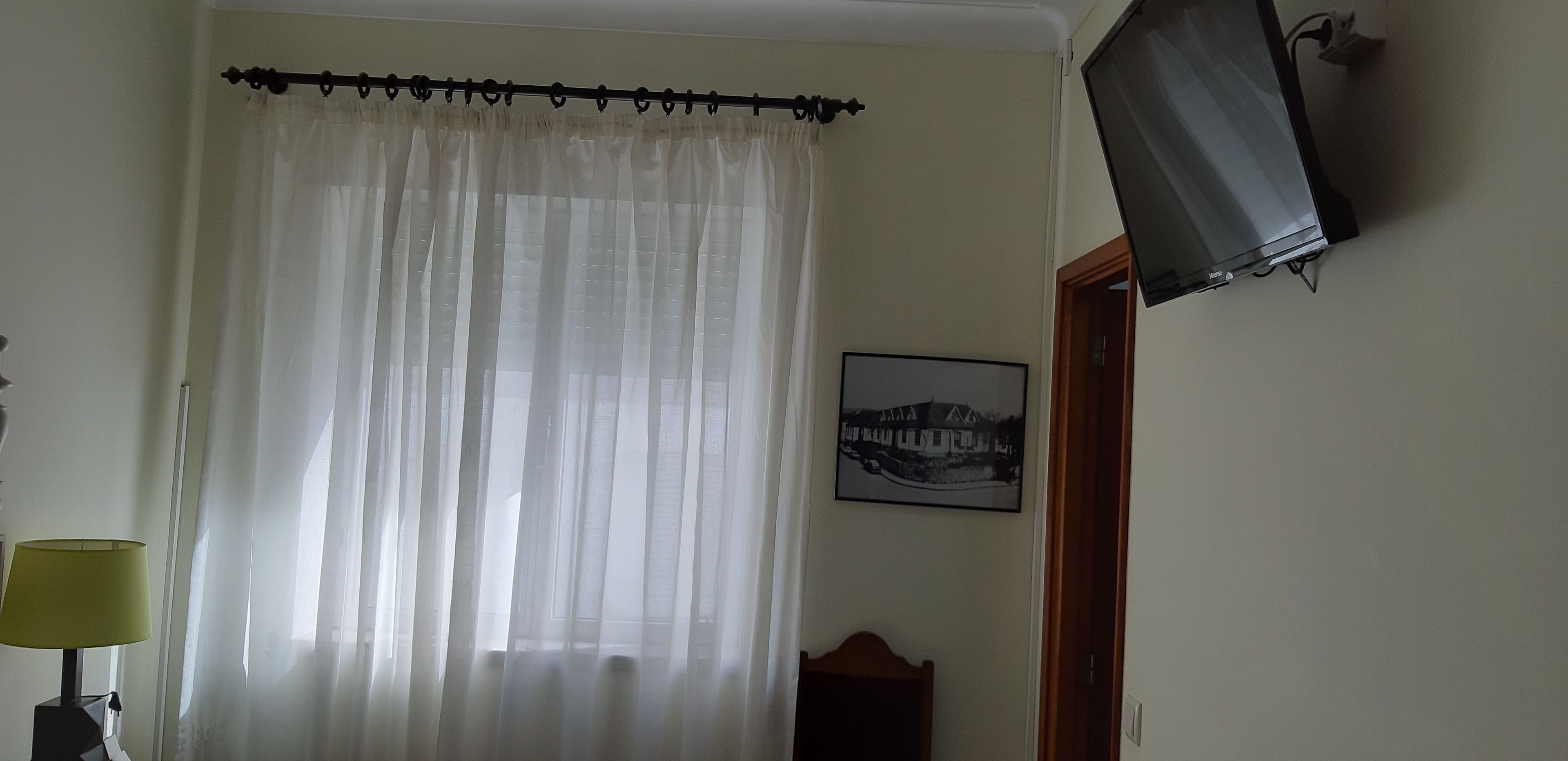 Una habitació amb una finestra amb cortines, una televisió penjada a la paret i un quadre emmarcat.