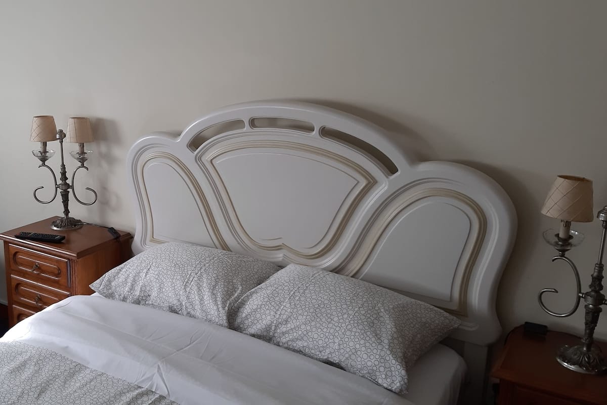 Een net opgemaakt bed met gestreept beddengoed, een houten nachtkastje met een lamp, een stoel met een groene kussens en een wandlamp.