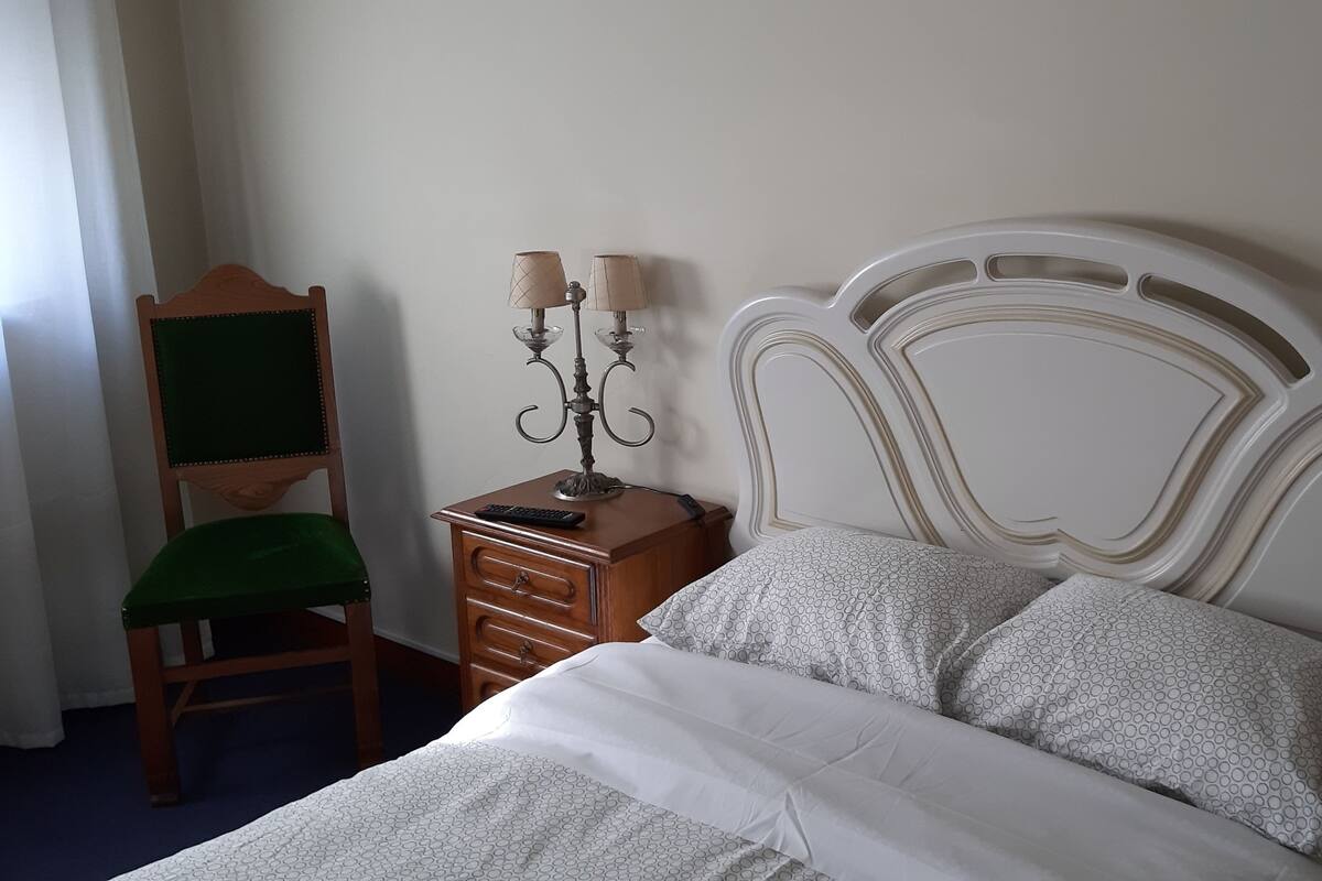 Een opgemaakt bed met een bedovertrek met patroon, een houten nachtkastje met een lamp, een stoel met een groene kussens en een wandlamp.