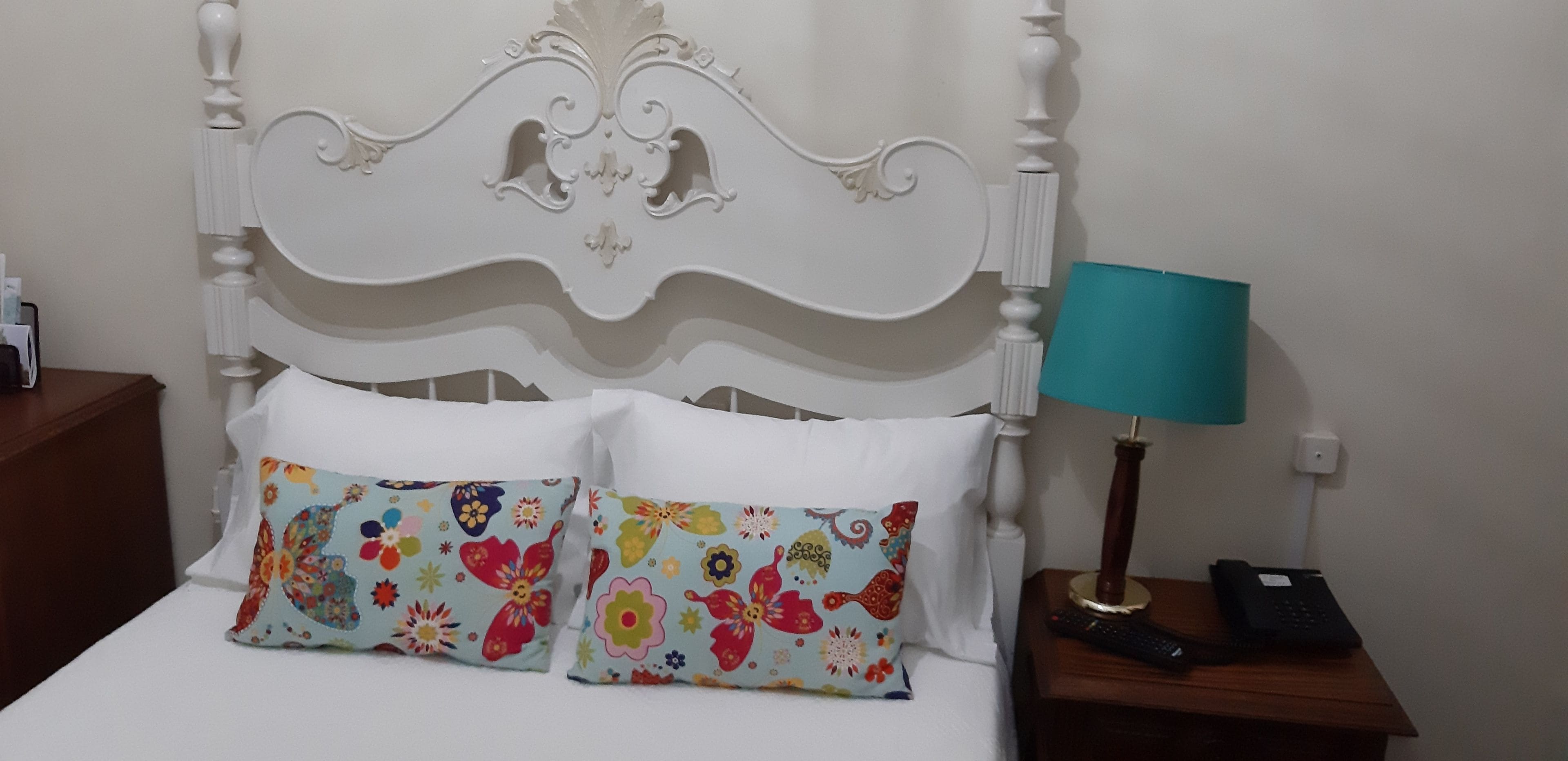 A bed with white bedding and colorful floral pillows, a white ornate headboard, a teal bedside lamp, and a bedside telephone.