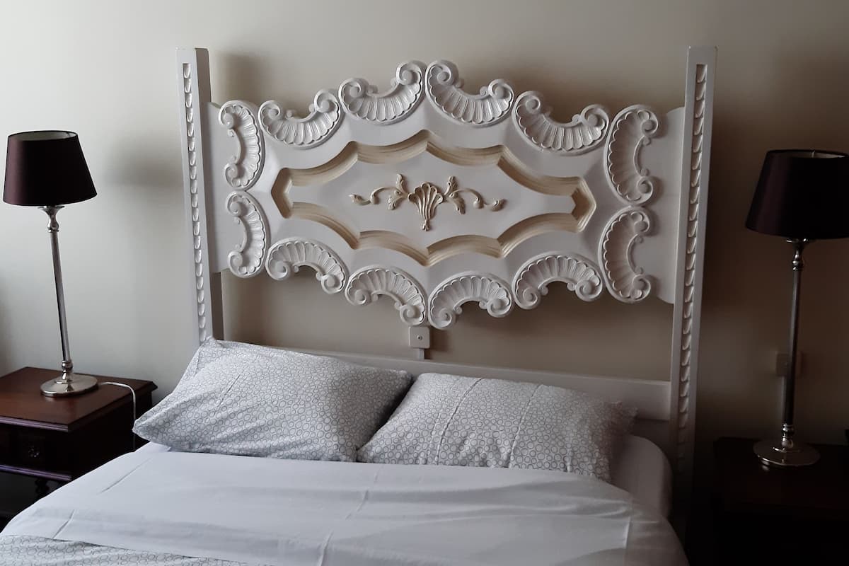 Een net opgemaakt bed met een decoratief hoofdbord, twee nachtkastjes met lampen en een houten stoel.