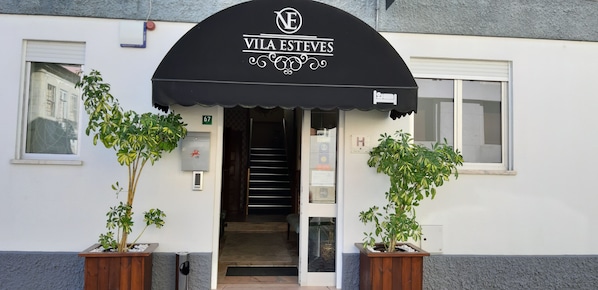 La entrada del hotel Vila Esteves con un toldo negro, una escalera que da al interior y dos plantas en macetas a cada lado.