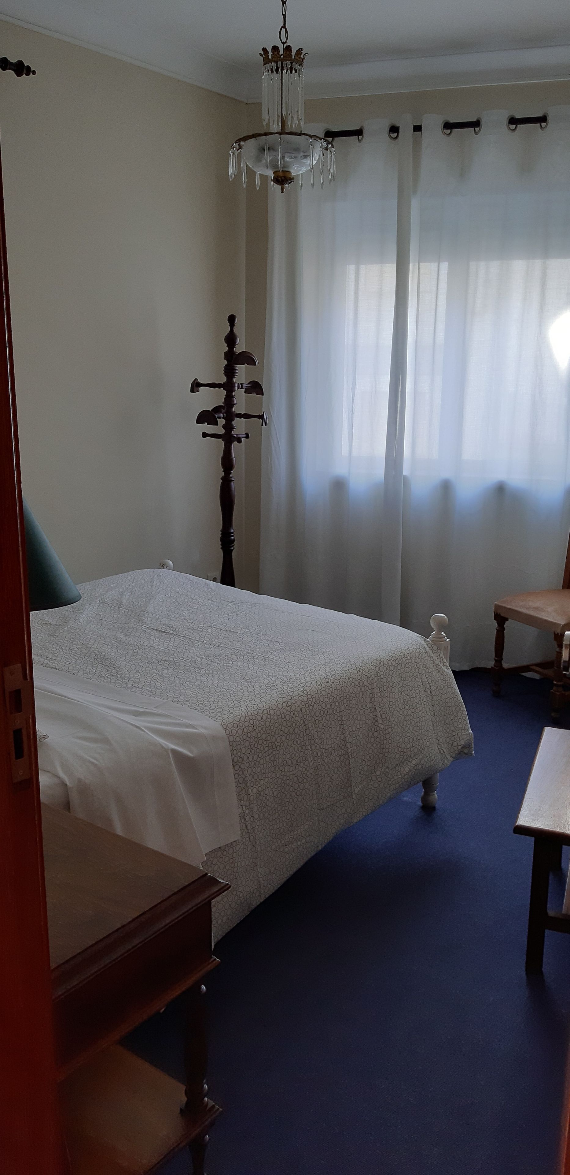 A bedroom with a large bed, a wooden wardrobe, a chandelier, and a window with sheer curtains.