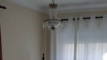 A bedroom with a large bed, a wooden wardrobe, a chandelier, and a window with sheer curtains.