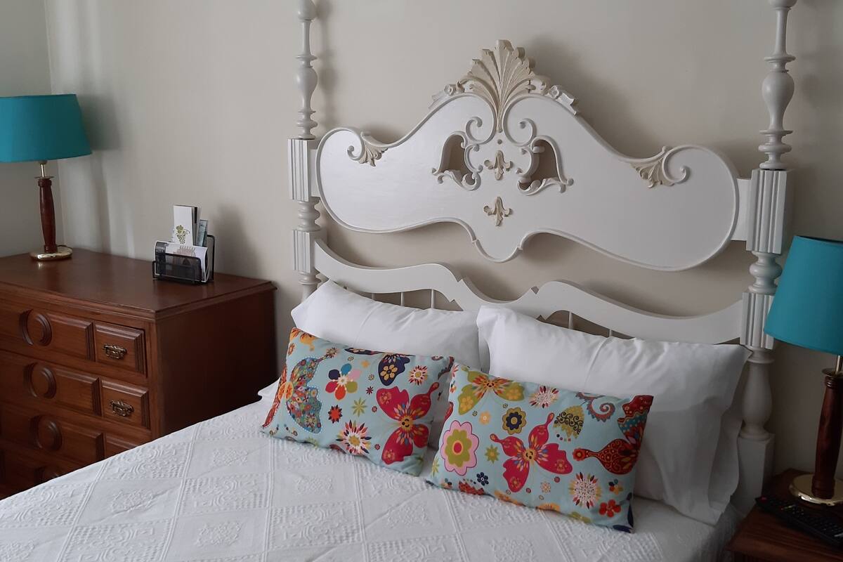 Een bed met een wit hoofdbord, twee bedlampen, een houten dressoir en een kleurrijk kussen.