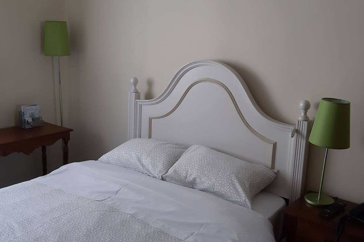 Een net opgemaakt bed met een wit hoofdeinde en bijpassende kussens, twee nachtkastjes met lampen, en een klein nachtkastje met een telefoon en afstandsbediening.