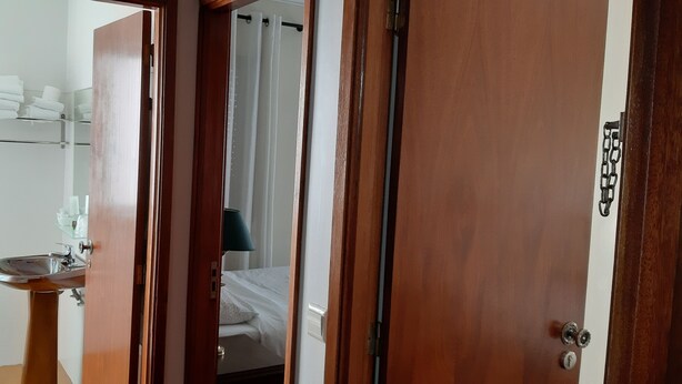 A hotel room with a wooden door, a bathroom with a sink and towel rack, and a bed with a white comforter.