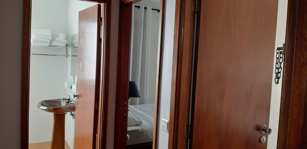 A hotel room with a wooden door, a bathroom with a sink and towel rack, and a bed with a white comforter. - Hotel Vila Esteves (Monção)