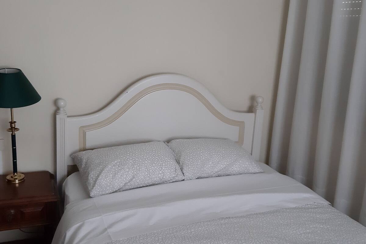 Een bed met een wit hoofdeinde en twee kussens, een nachtkastje met een lamp en een raam met gordijnen.