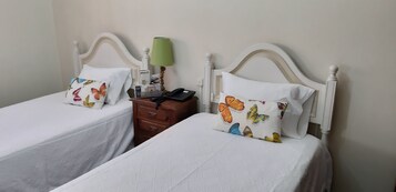 Dos camas individuales con ropa de cama blanca y almohadas de mariposas de colores, una mesita de noche de madera con una lámpara verde y un teléfono negro.