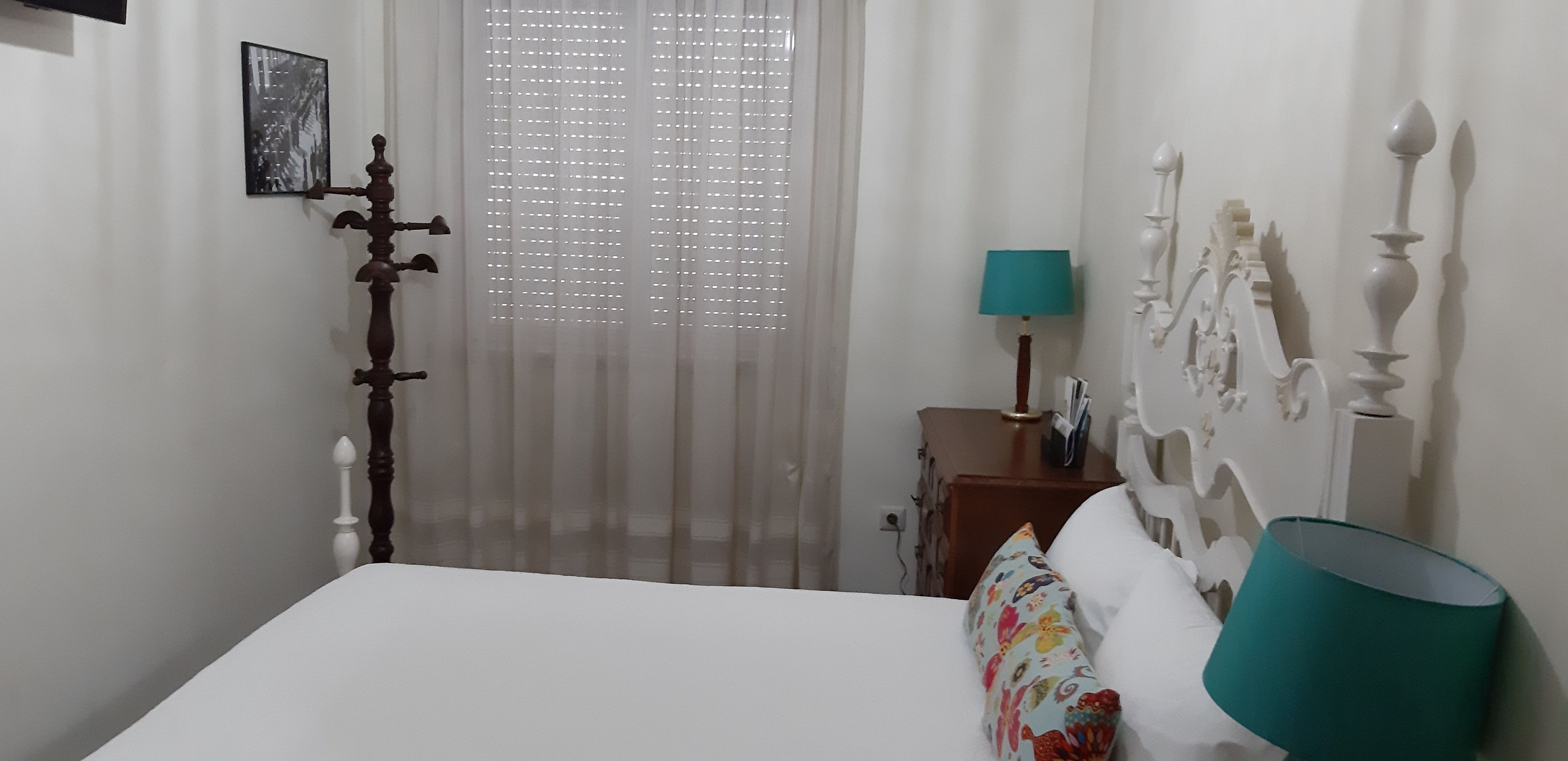 A bedroom with a bed, a nightstand, a lamp, a window with curtains, and a wall-mounted shelf.