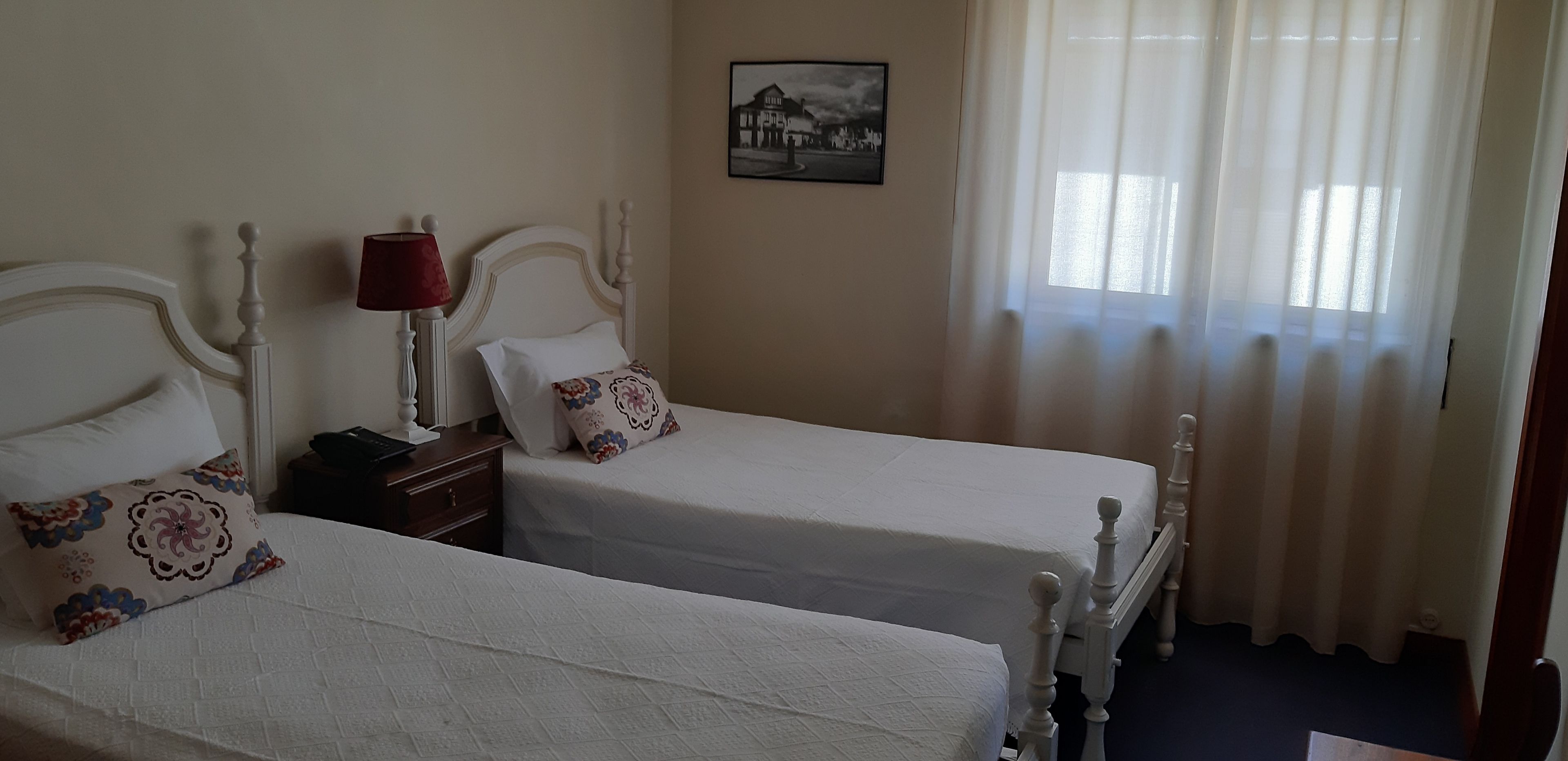 A hotel room with two single beds, a nightstand with a lamp, a framed picture on the wall, and a window with sheer curtains.