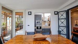 Et spiseområde med et stort træbord, stole og en dekorativ skål som centerpiece. Et køkken er synligt i baggrunden med moderne hårde hvidevarer.