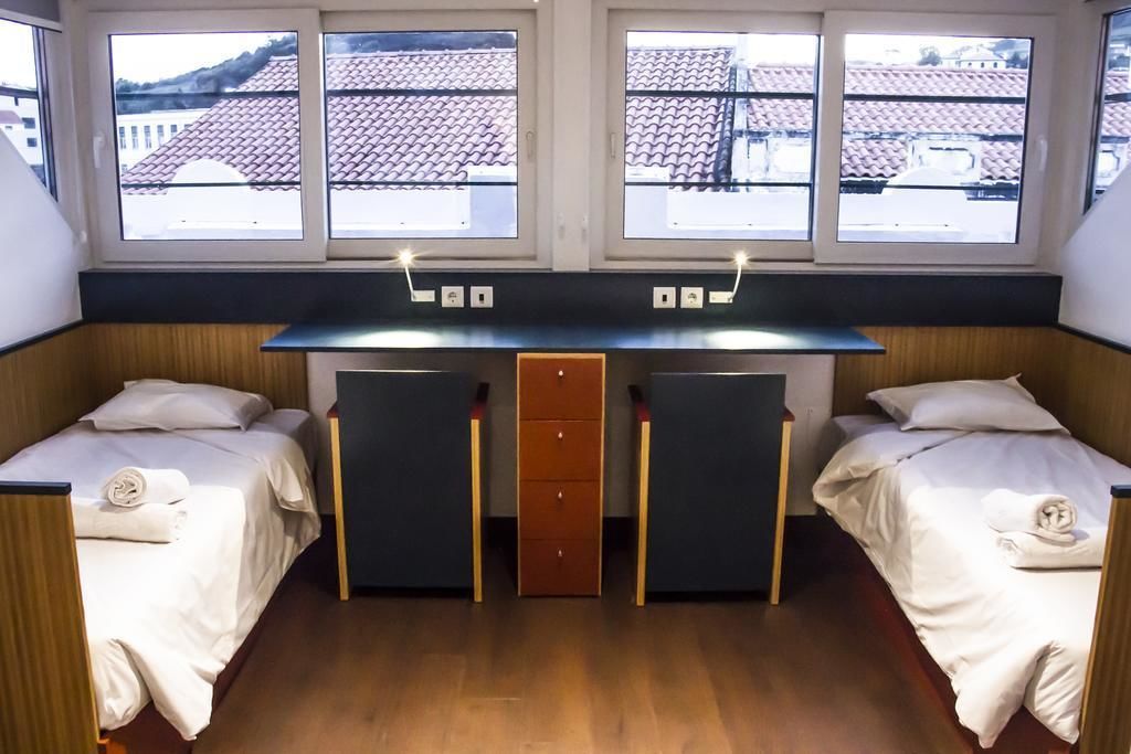 A room with two beds, a desk, and a view of buildings through large windows.