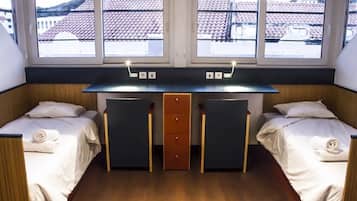 A room with two beds, a desk, and a view of buildings through large windows.