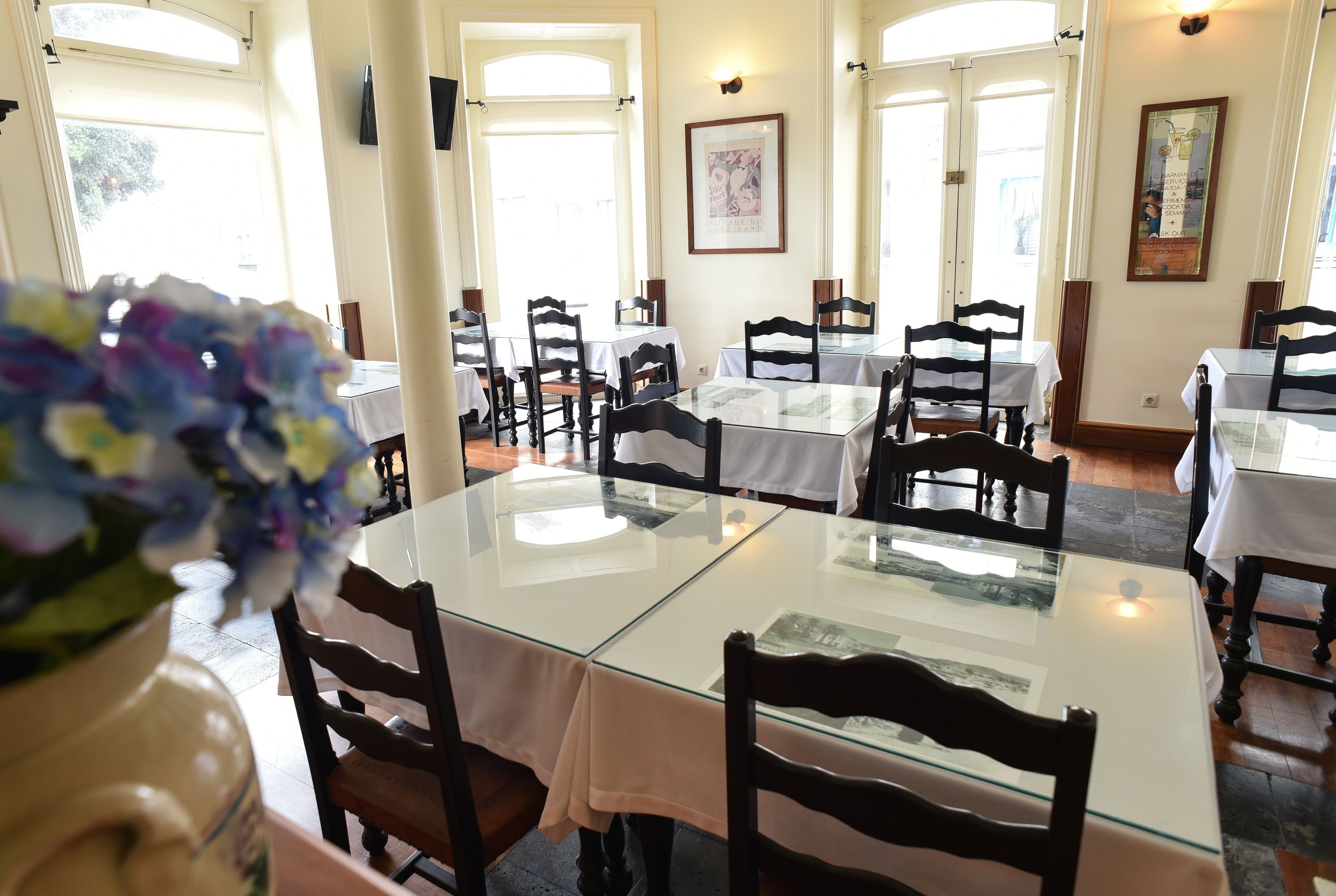A dining area with glass-top tables and wooden chairs, framed artwork on the walls, and large windows allowing natural light.