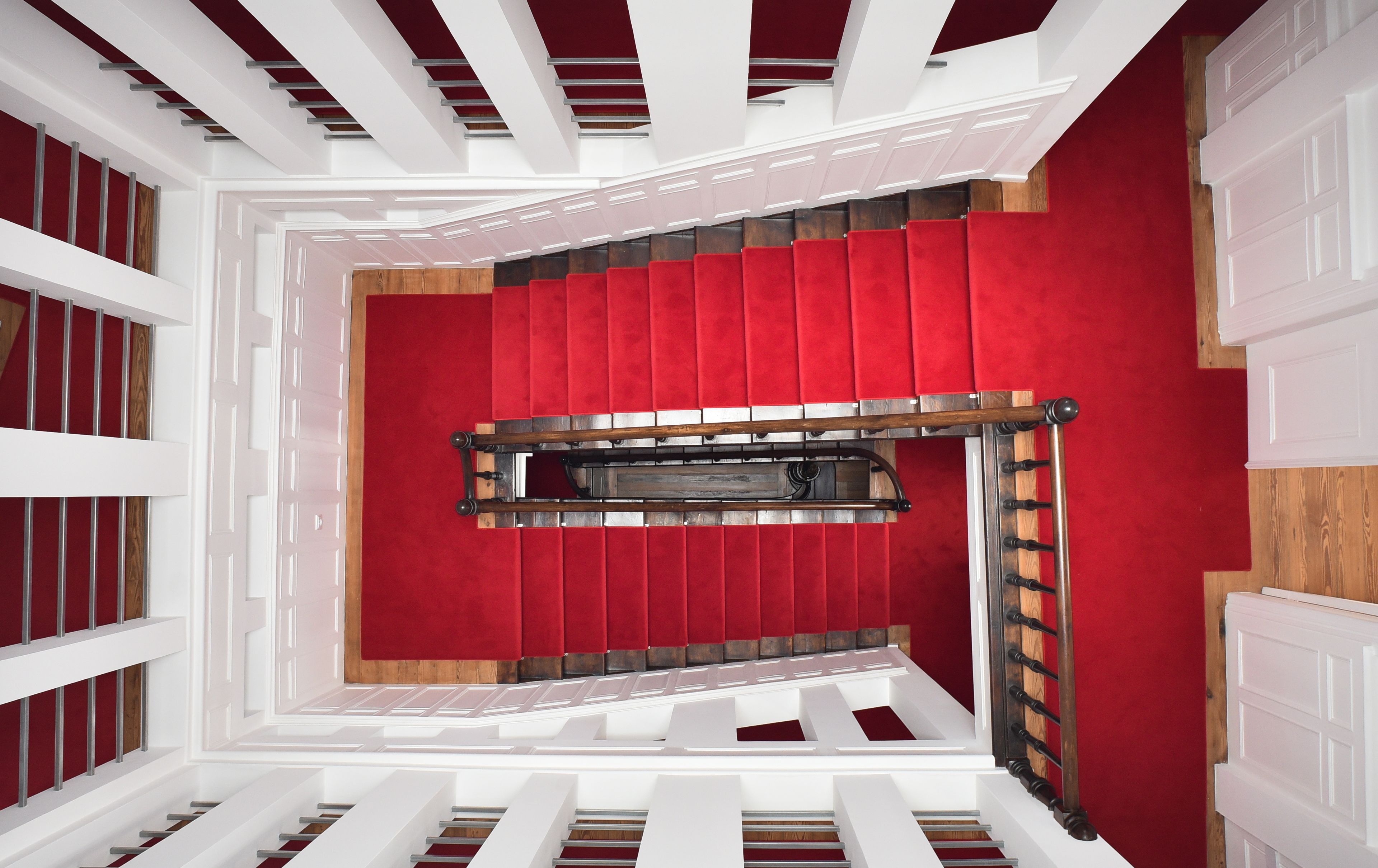 Una escalera con un muro cubierto de alfombra roja, barandillas blancas y un pasamanos de madera.