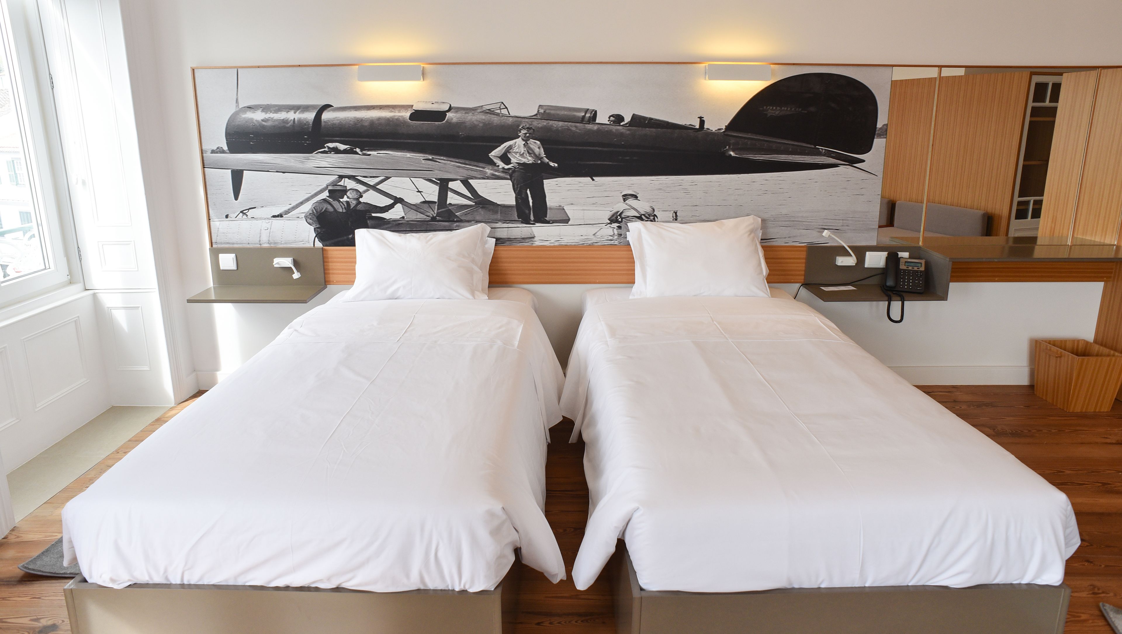 A hotel room with two single beds, a large framed picture of an airplane on the wall, a bedside table with a lamp, and a wooden floor.
