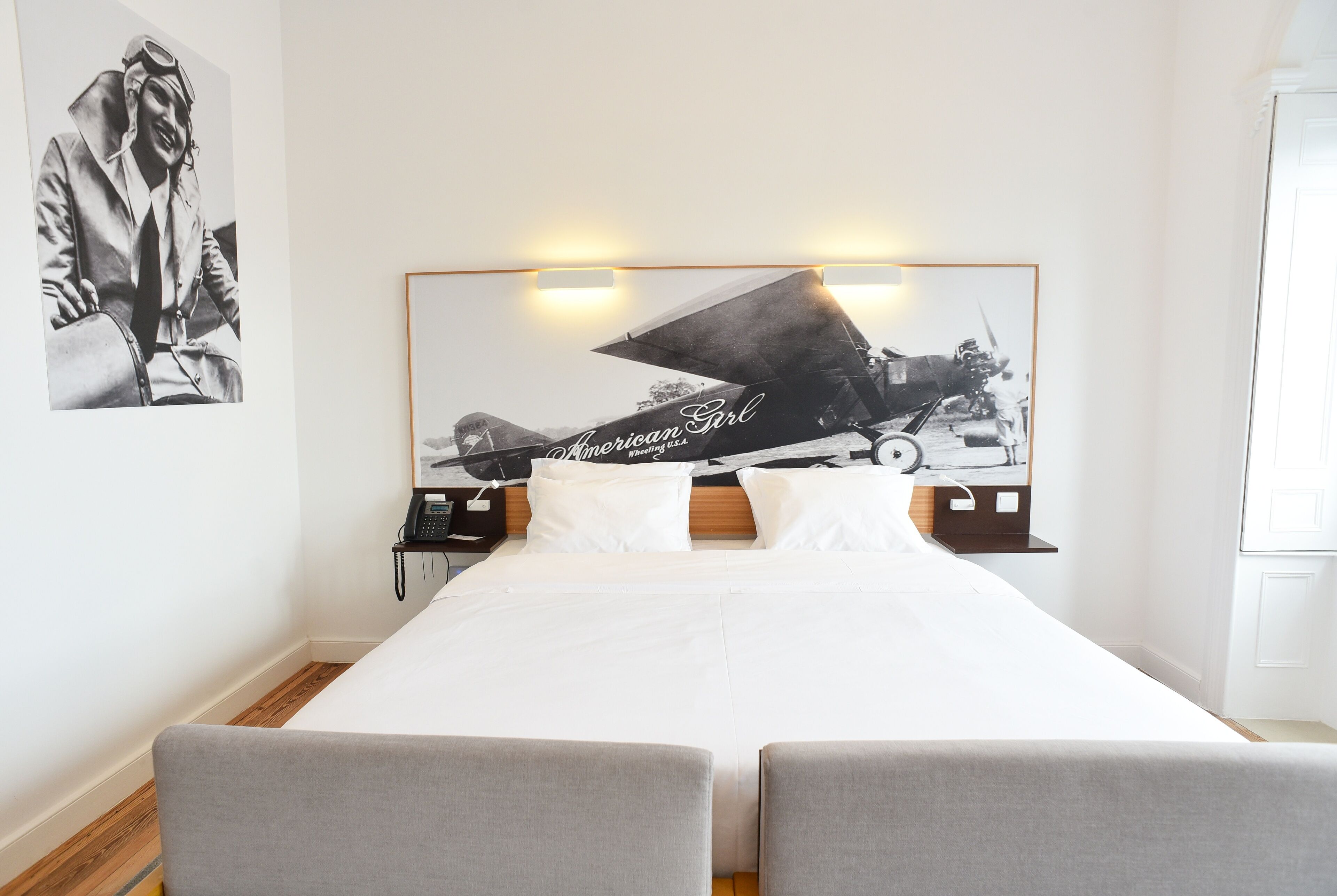 A modern hotel room with a large bed, two grey armchairs, a headboard with a vintage airplane photo, and a small bedside table with a lamp.