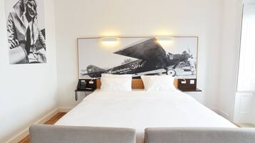 A modern hotel room with a large bed, two grey armchairs, a headboard with a vintage airplane photo, and a small bedside table with a lamp.