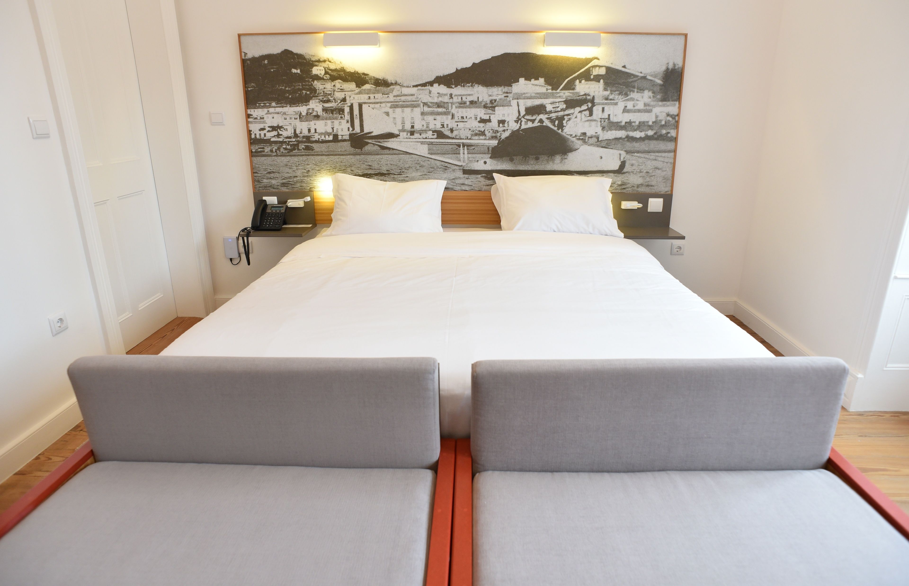 Une chambre d’hôtel avec un lit, deux tables de chevet, une petite table et un grand tableau accroché au mur représentant une ville côtière.
