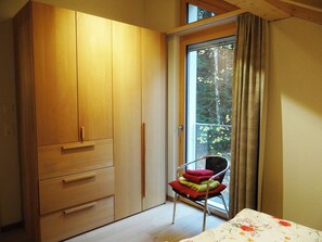 Une chambre avec une armoire en bois, un fauteuil avec des coussins, un lit recouvert d’une couvre-lit à motifs floraux et une fenêtre donnant sur des arbres.