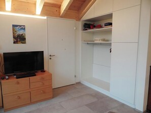 A room with a television on a wooden cabinet, a white wardrobe, and a shelf with various items.