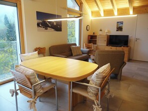 A cozy living room with a wooden dining table, comfortable seating, a television, and a large window offering a view of the outdoors.