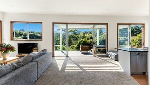 Una sala moderna con un sofá gris grande, un televisor de pantalla plana y una planta en maceta. Hay una puerta corrediza de vidrio que da a un patio exterior con una mesa y sillas de madera.