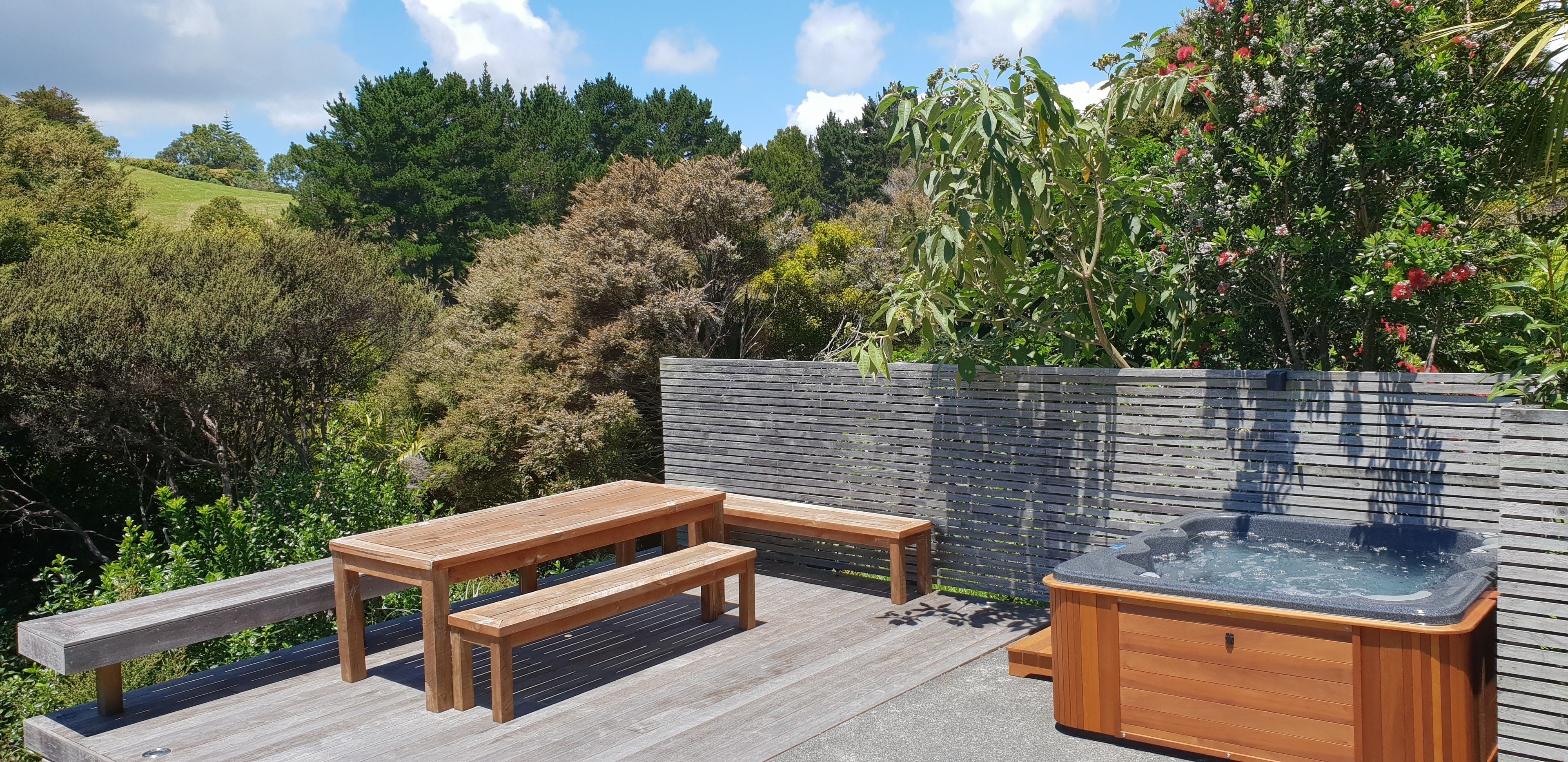 A wooden deck with a hot tub and benches, surrounded by lush greenery and a wooden fence.