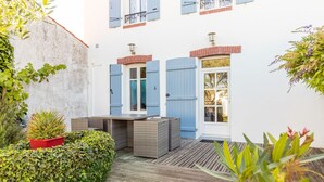 Un patio con una terraza de madera, una maceta roja con una planta y un edificio blanco con contraventanas azules y una puerta de cristal.