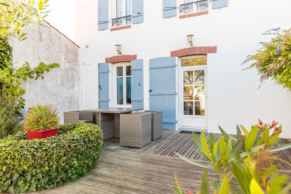 A courtyard with a wooden deck, a red planter with a plant, and a white building with blue shutters and a glass door.