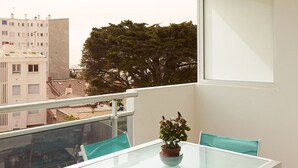 Un balcón con una mesa de vidrio y sillas turquesas, una planta en maceta y vistas a edificios y árboles.