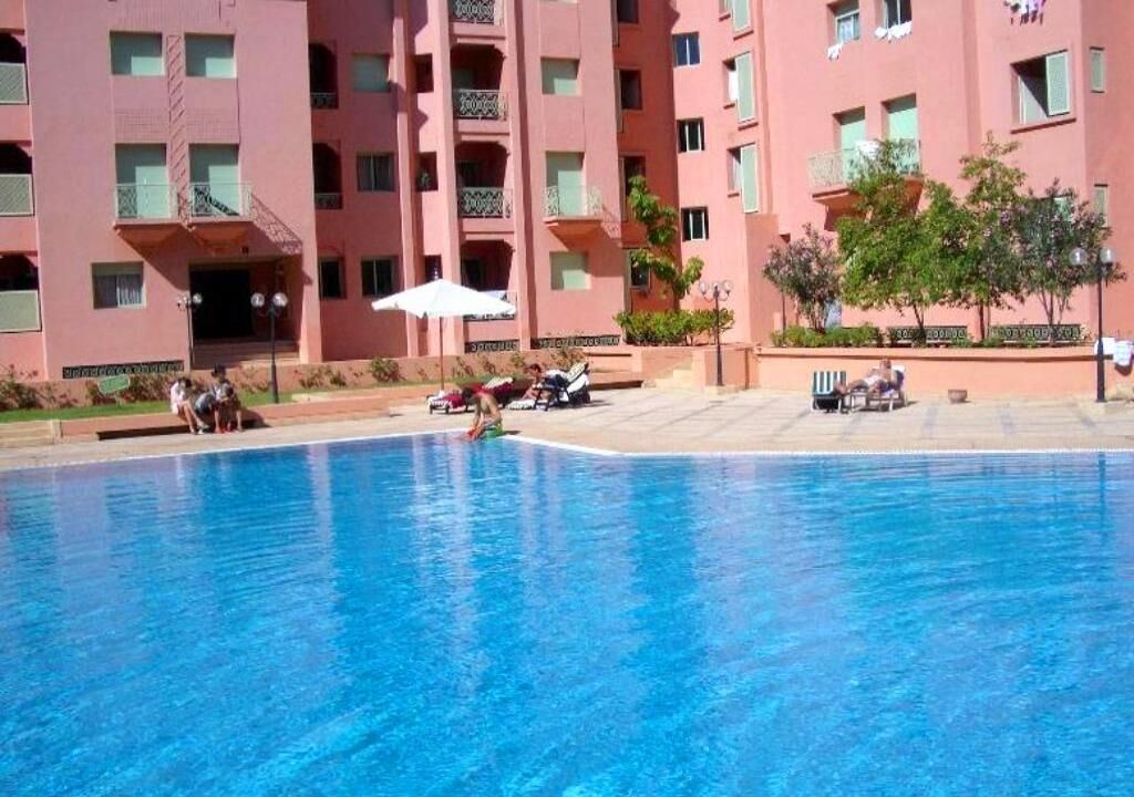 A swimming pool with a pink building in the background, a few people near the pool, and some umbrellas.