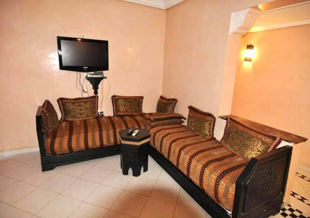 A room with a sofa bed, a small table, a television, and a wall-mounted light fixture.