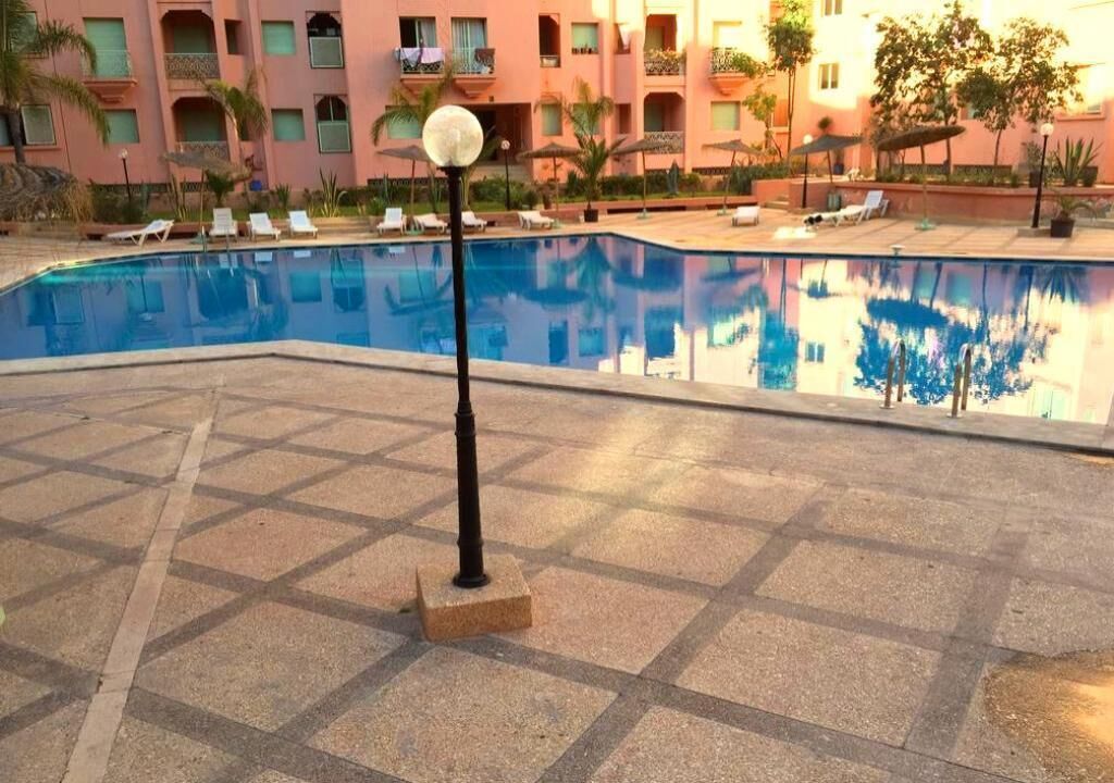 A swimming pool with lounge chairs and umbrellas, surrounded by a tiled patio and a building with balconies.
