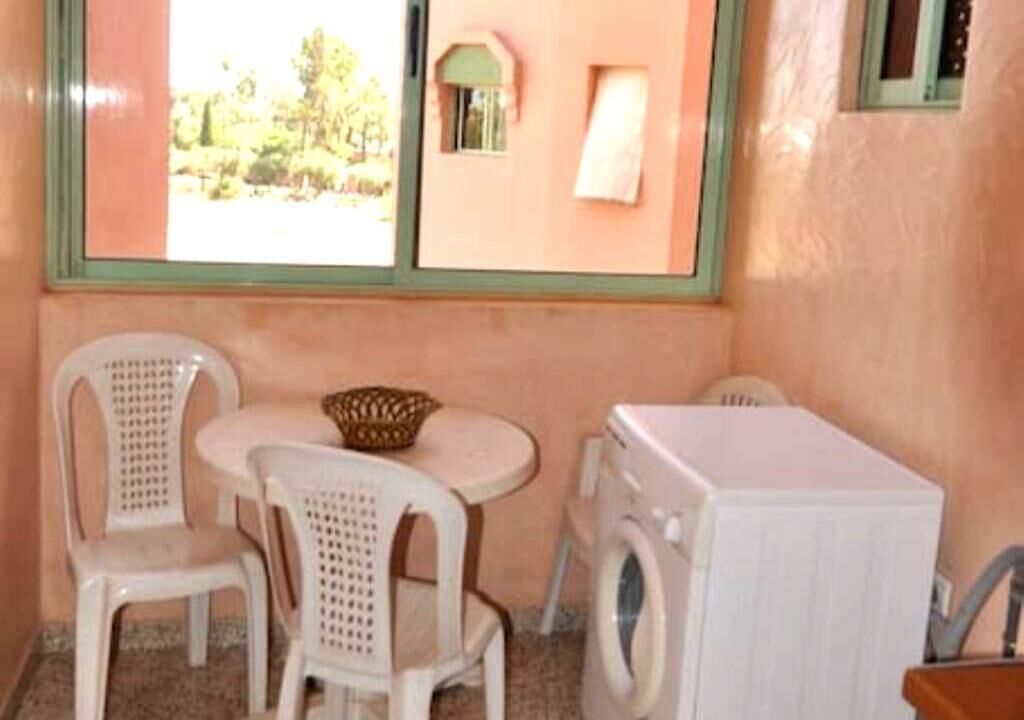 Een kleine kamer met een wasmachine, twee witte plastic stoelen, een ronde tafel en een raam met uitzicht op groen.