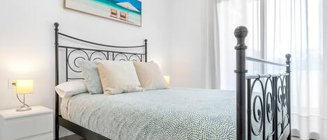 Un lit bien fait, recouvert d’une couvre-lit à motifs, une table de chevet avec une lampe et un tableau représentant une scène de plage accroché au mur.