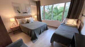 A hotel room with two beds, a nightstand, a lamp, and a window with a view of greenery. - Cabarete Beach Front Houses A8.  TOP LOCATION IN CABARETE (Cabarete)