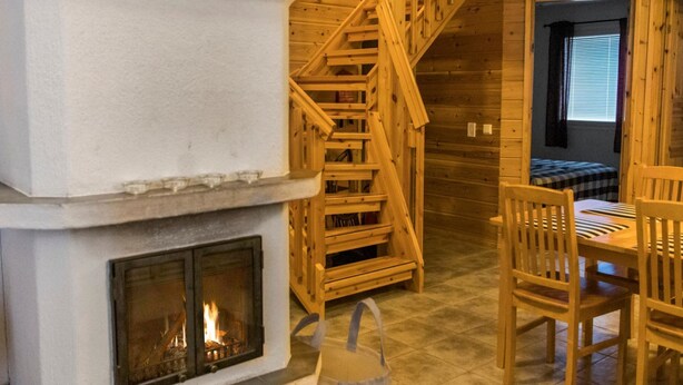 Interior acollidor d'una cabana amb una llar de foc, unes escales de fusta i una zona de menjador.