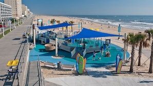 Un terrain de jeux au bord de la plage, doté de structures de protection solaire bleues, d’une piscine et de décorations en forme de planche de surf.