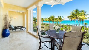A balcony with a table and chairs, overlooking a beach with palm trees and the ocean.