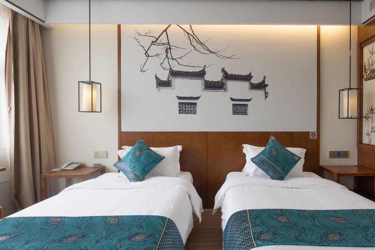A hotel room with two beds, each with a teal-patterned bedspread, a wooden headboard, and a wall-mounted artwork depicting traditional architecture.