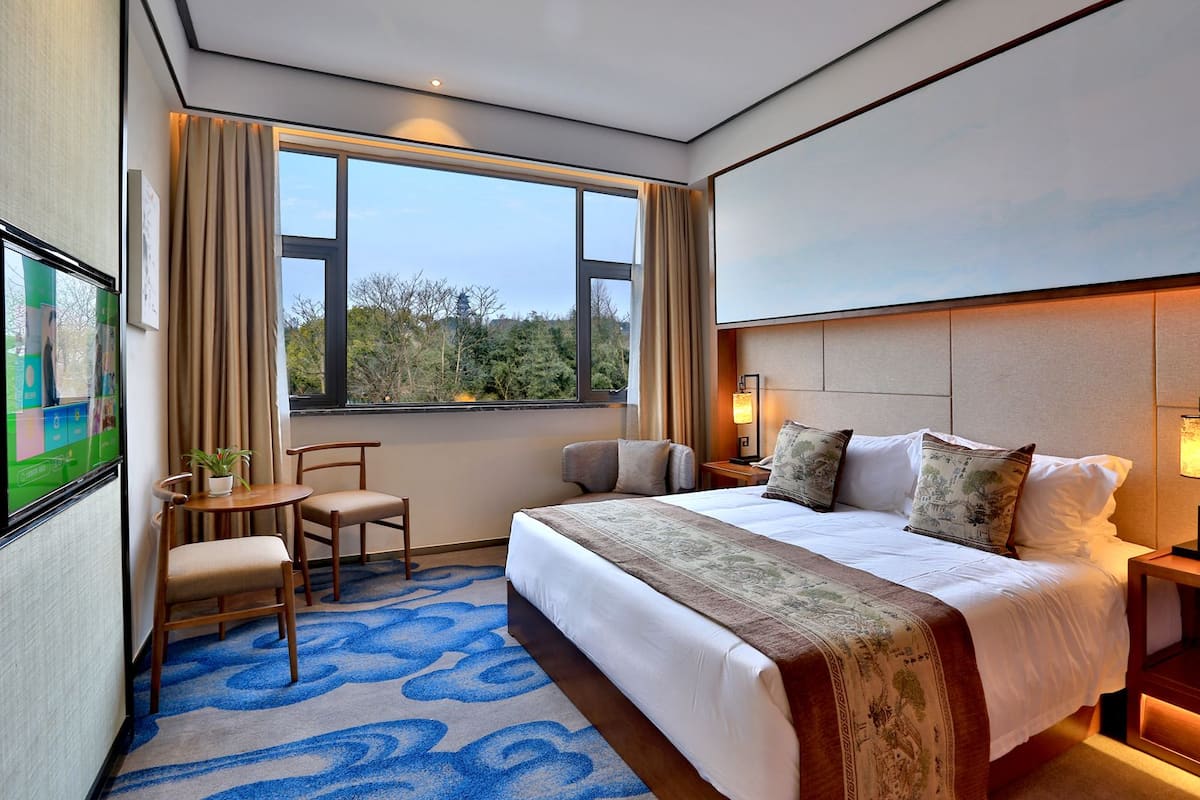 A hotel room with a large bed, a desk with a chair, a small table with a plant, and a large window with curtains.