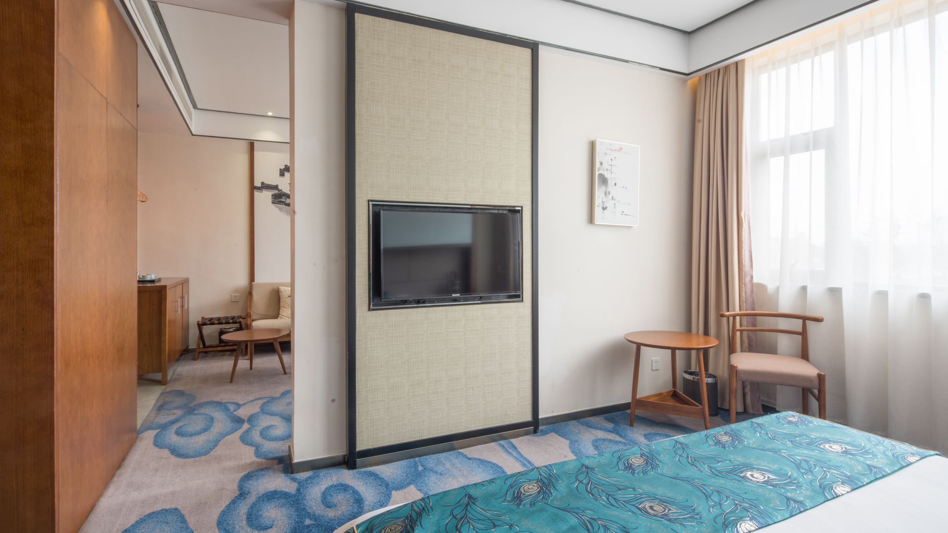 A hotel room with a sliding door, a TV mounted on the wall, a small round table, two chairs, a bed with a blue patterned bedspread, and a window with curtains.