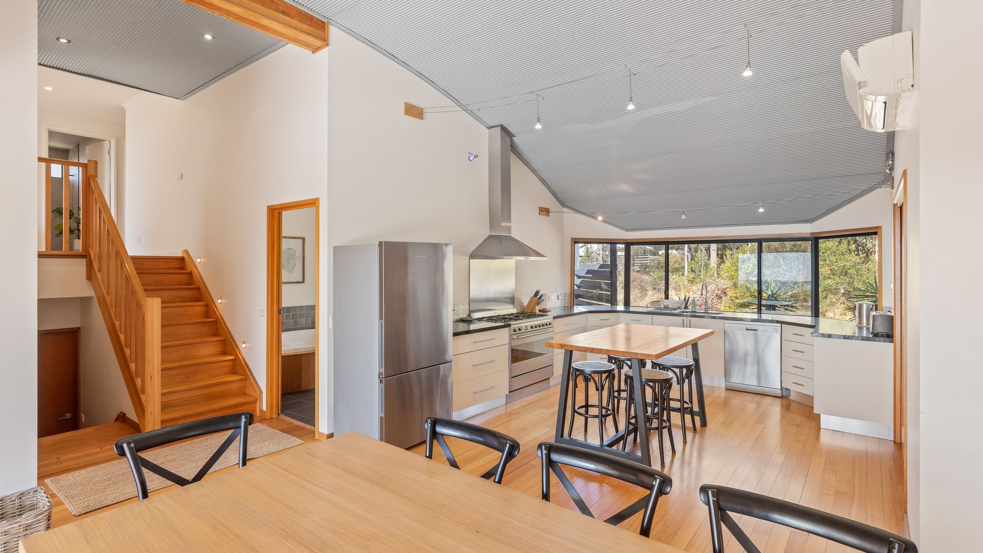 A modern kitchen with a dining area, wooden staircase, and large windows.